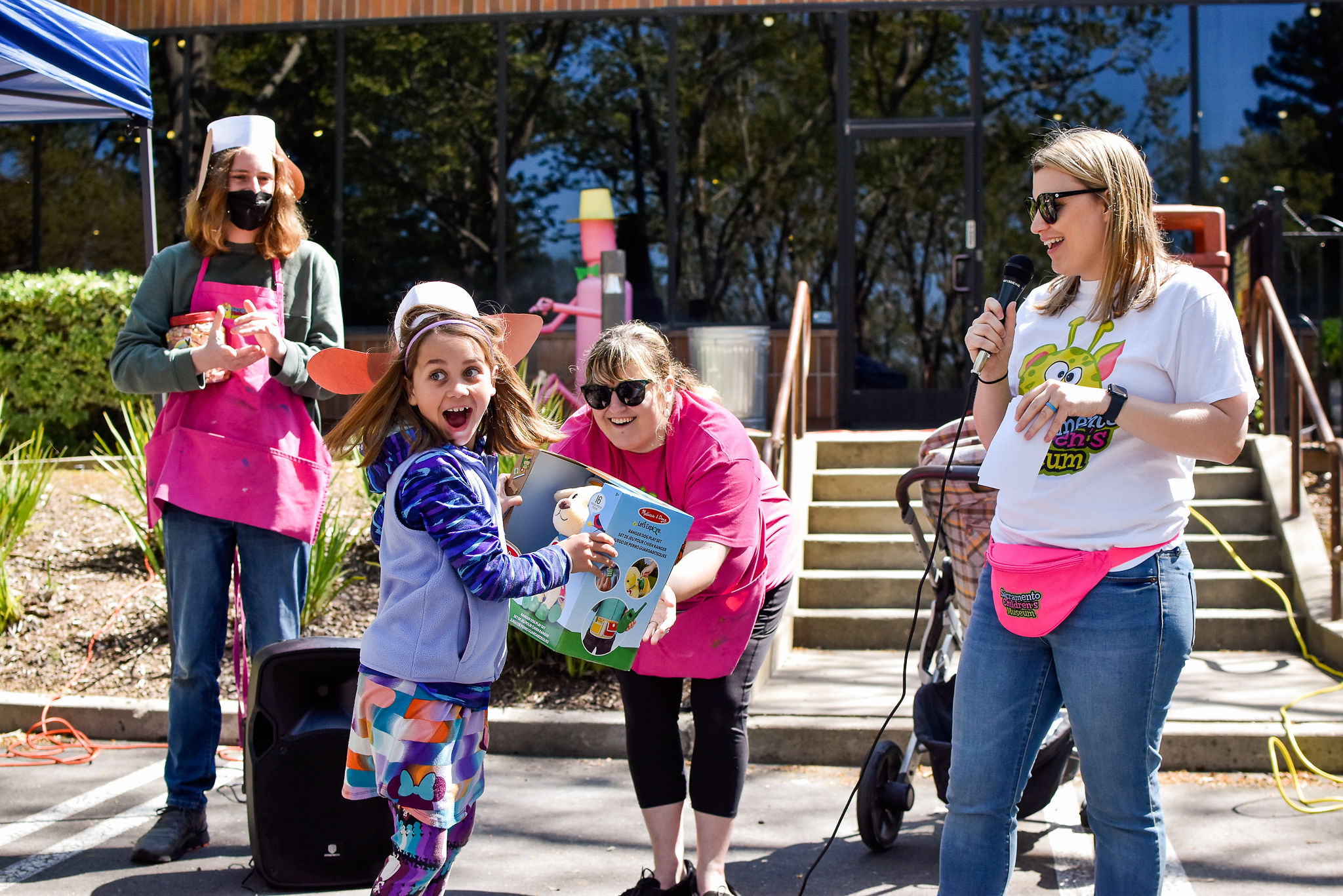 Three museum workers give a prize to a very excited little girl wearing a handcrafted hat