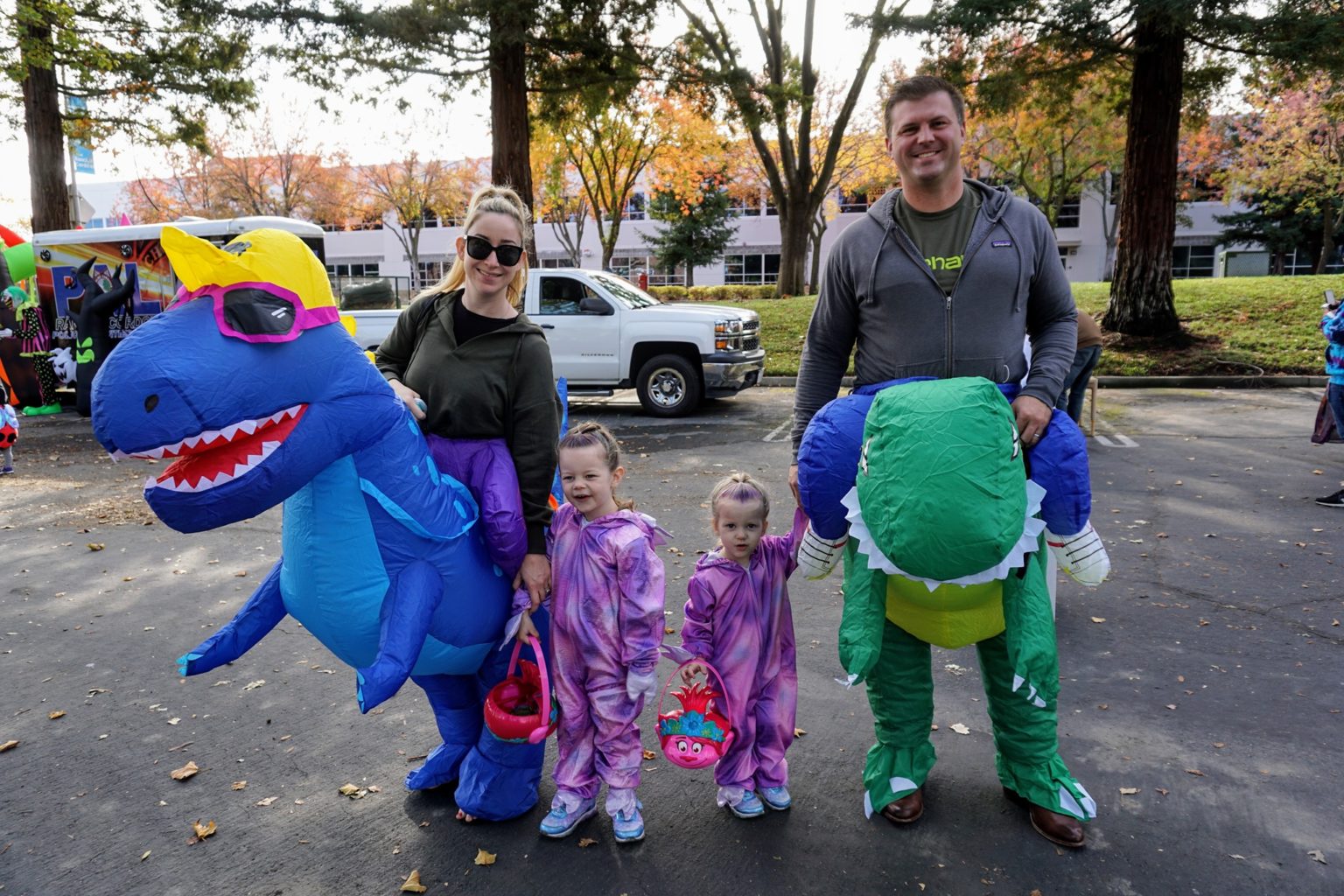 A mother and father dressed in blow up dinosaur rider costumes hold the hands of two little girls dressed in purple dinosaur onesies