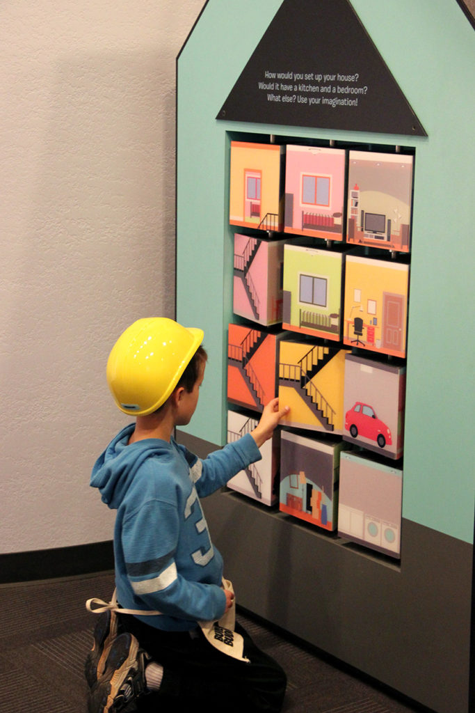 A Boy In A Hard Hat Kneels In Front Of A Wall Of Revolving Squares That Make Up Rooms In A House
