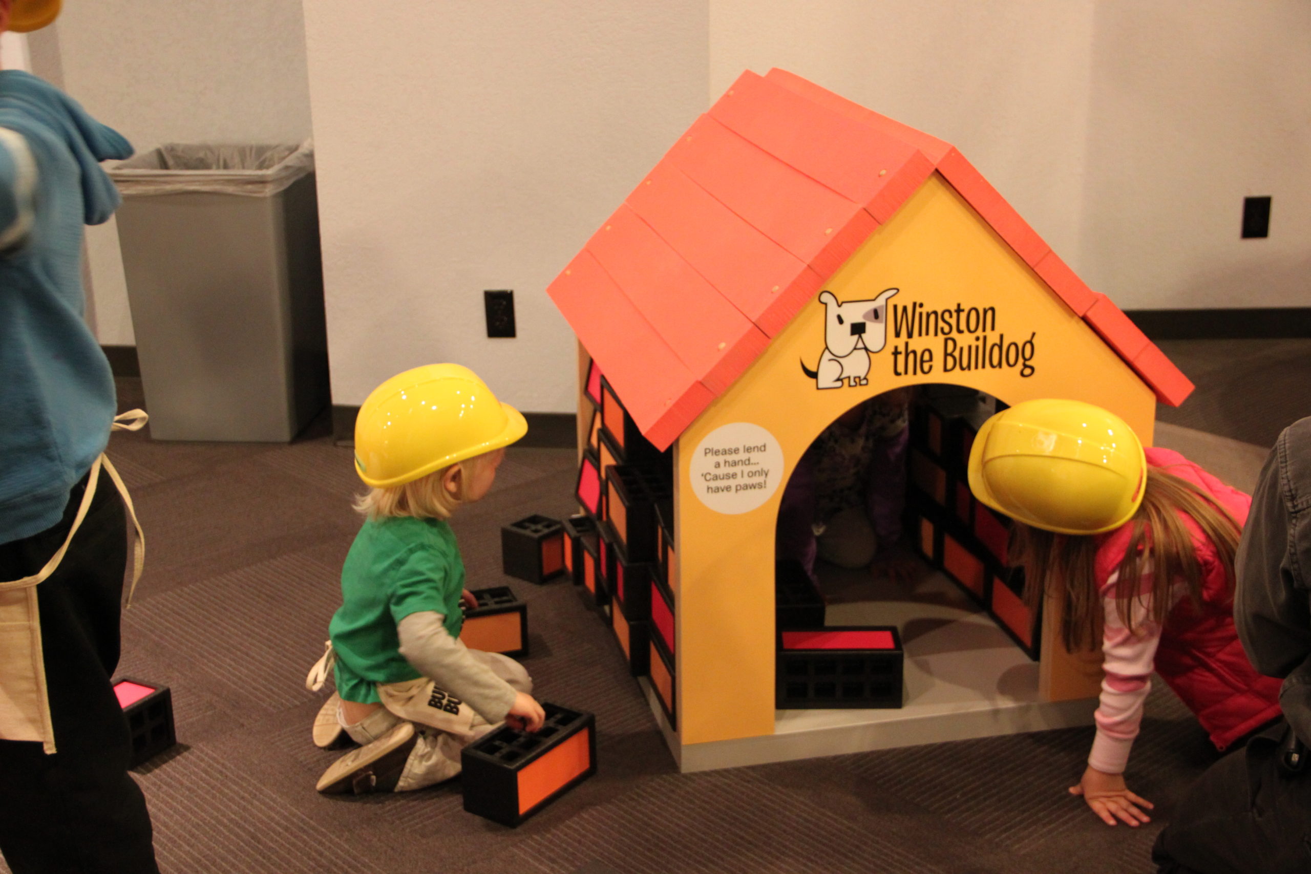 Two children in yellow hard hats use building blocks to build an orange dog house