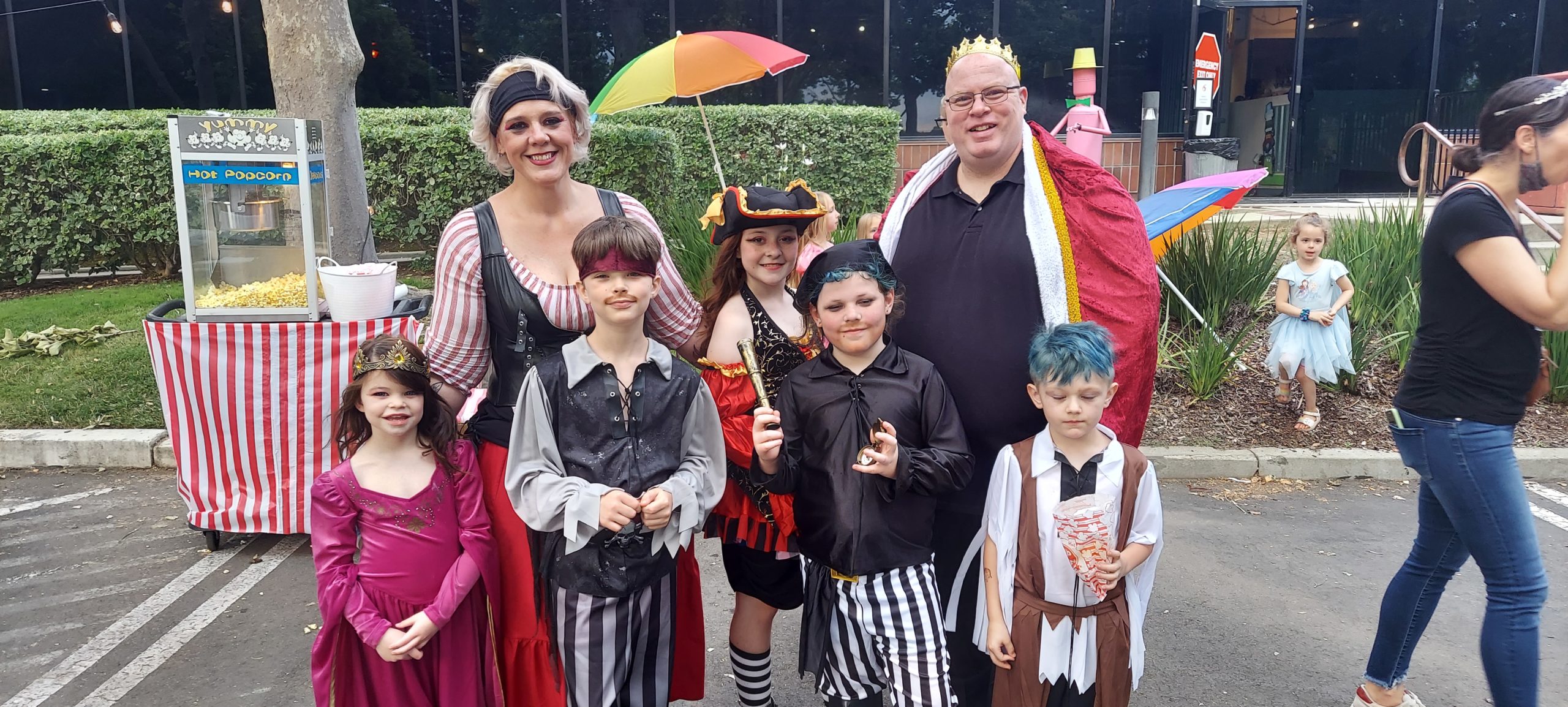 A family of 7 dressed as pirates and princesses pose outside in front of a popcorn cart