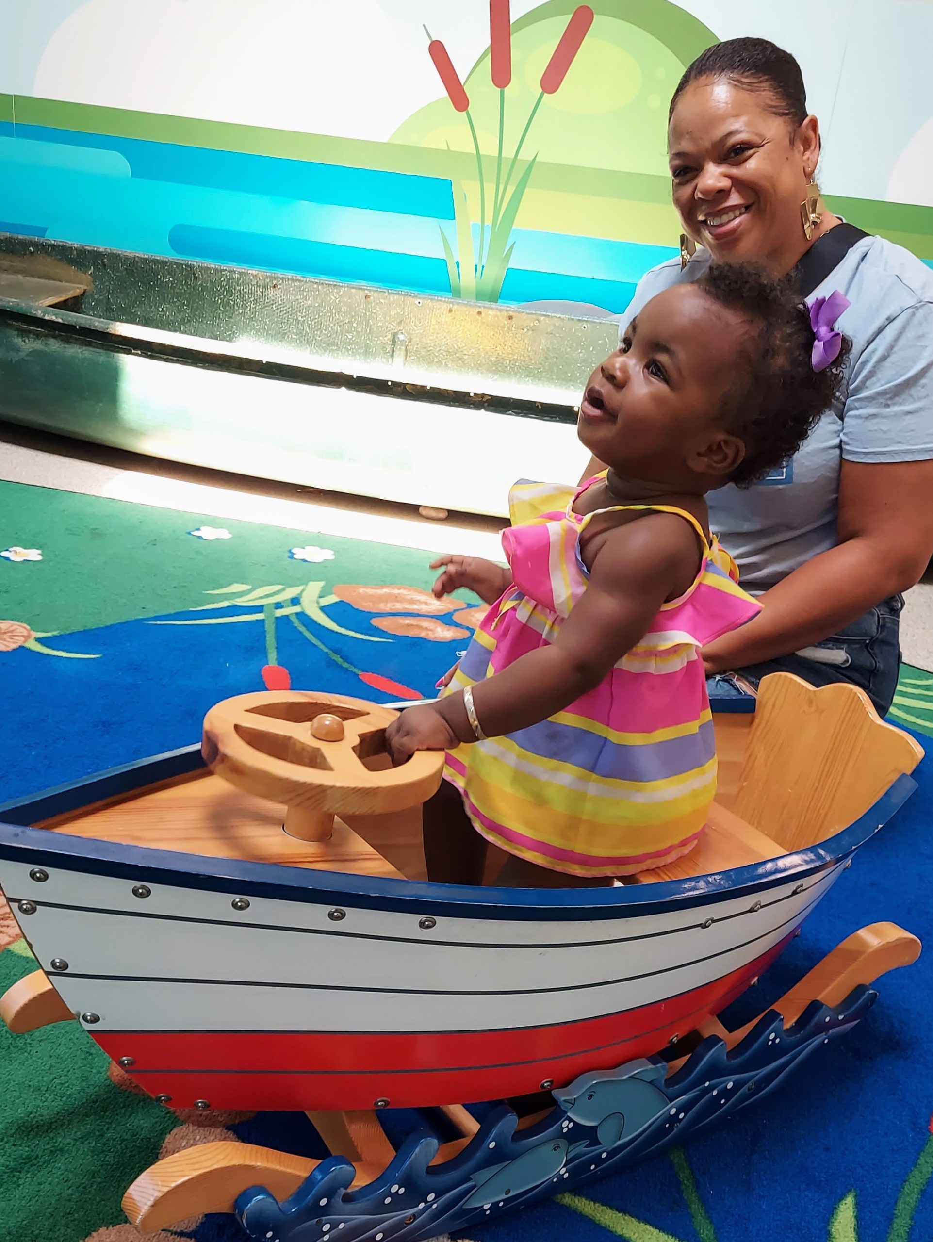 A baby smiles while riding a rocking boat toy in Camp Rancho, with her mother smiling in the background
