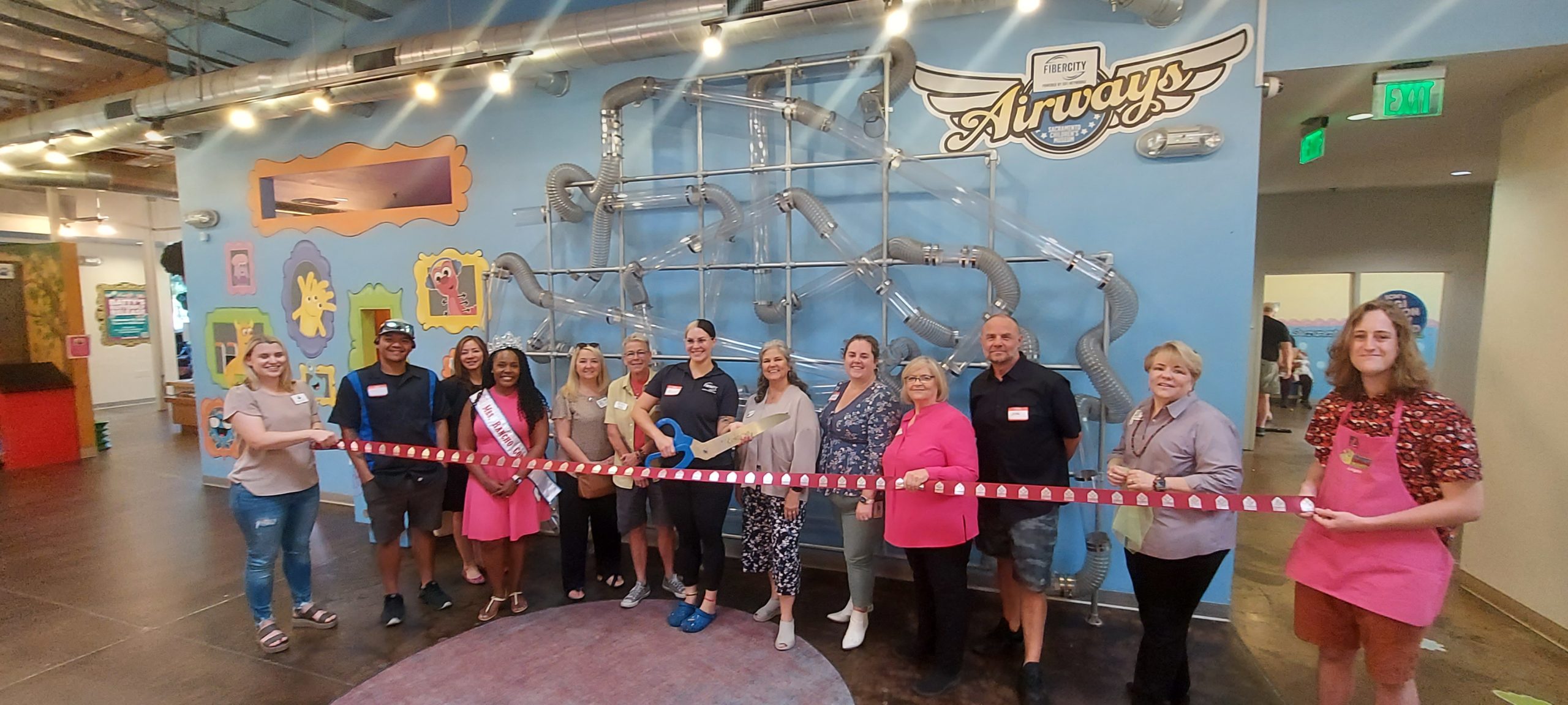 A line of people holding an opening ribbon poses in front of the Airways play exhibit