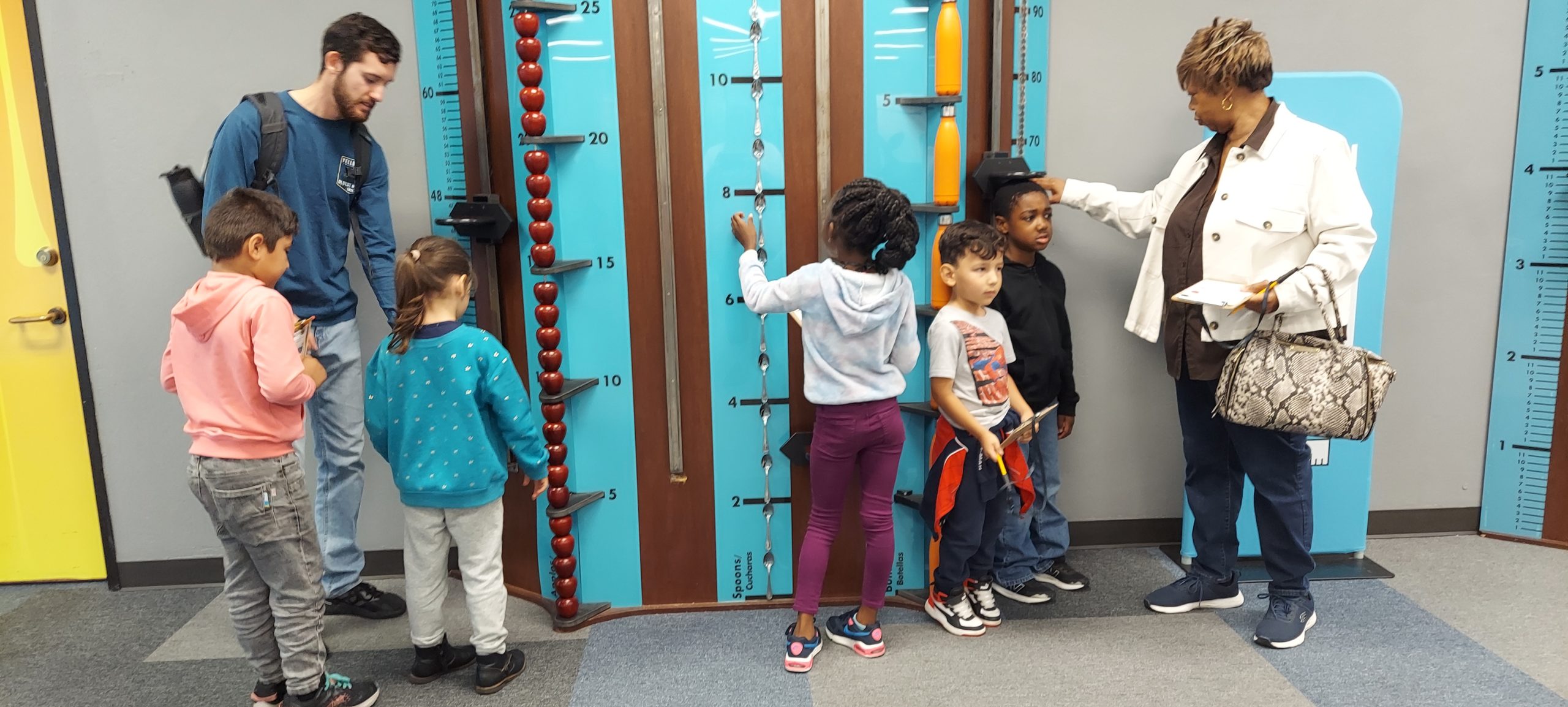Five children wait for their turn to be measured on a Measurement Rules exhibit piece, helped by two parent chaperones.