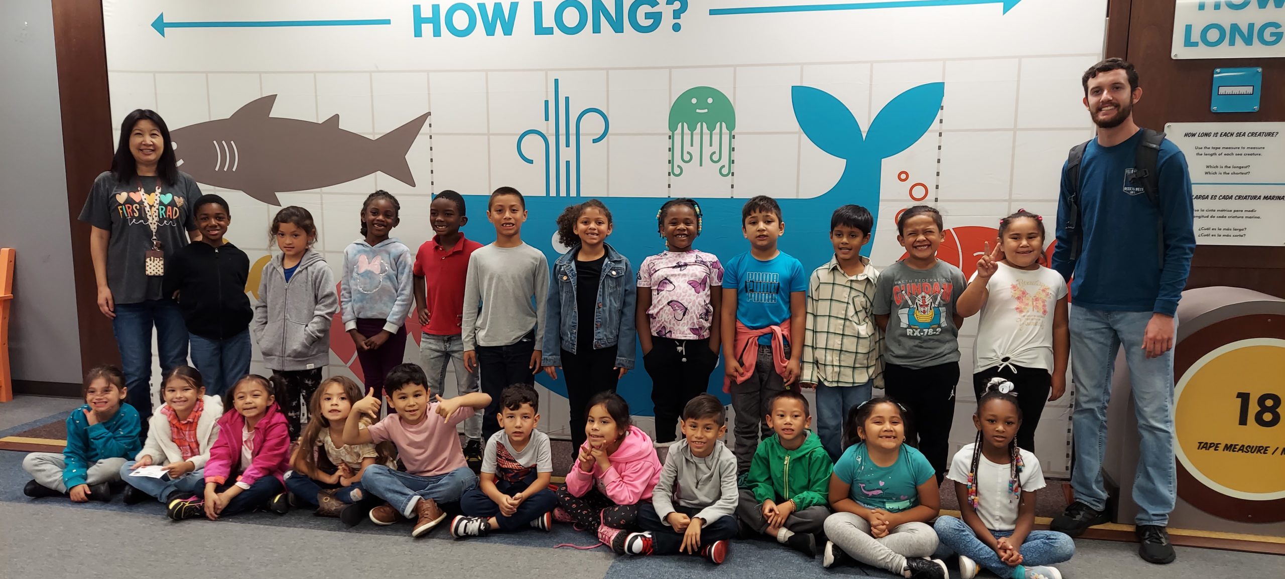 Two lines of students and their 2 teachers smile in a field trip photograph in front of a Measurement Rules exhibit piece.