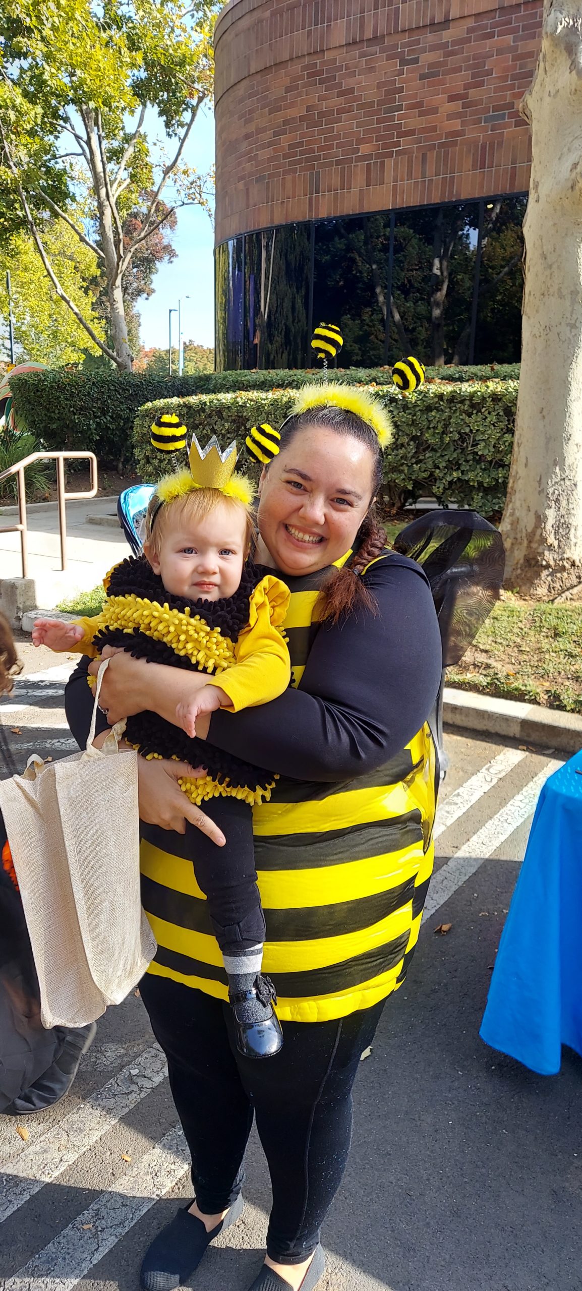 A mother dressed as a bumblebee smiles while holding her baby, dressed as a queen bumblebee.