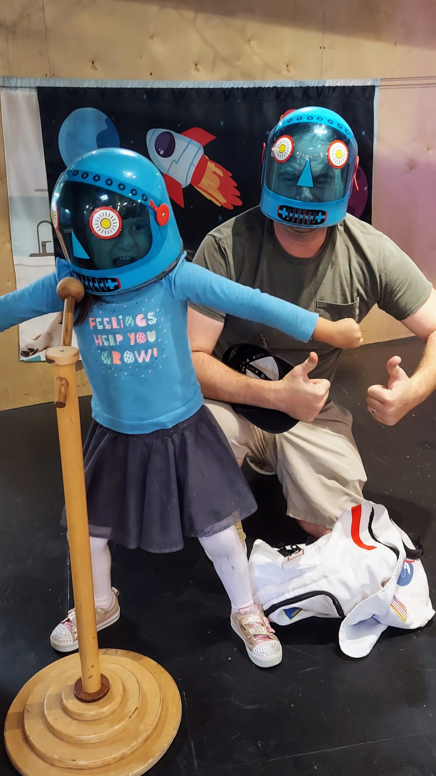 A daughter and dad, dressed in blue astronaut helmets, pose together in front of a wooden microphone.