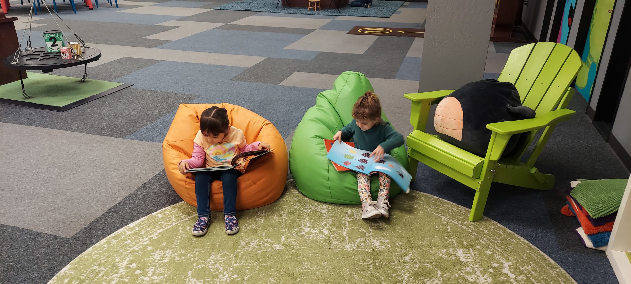 Two young girls read books while sitting on beanbag chairs next to a large green folding chair