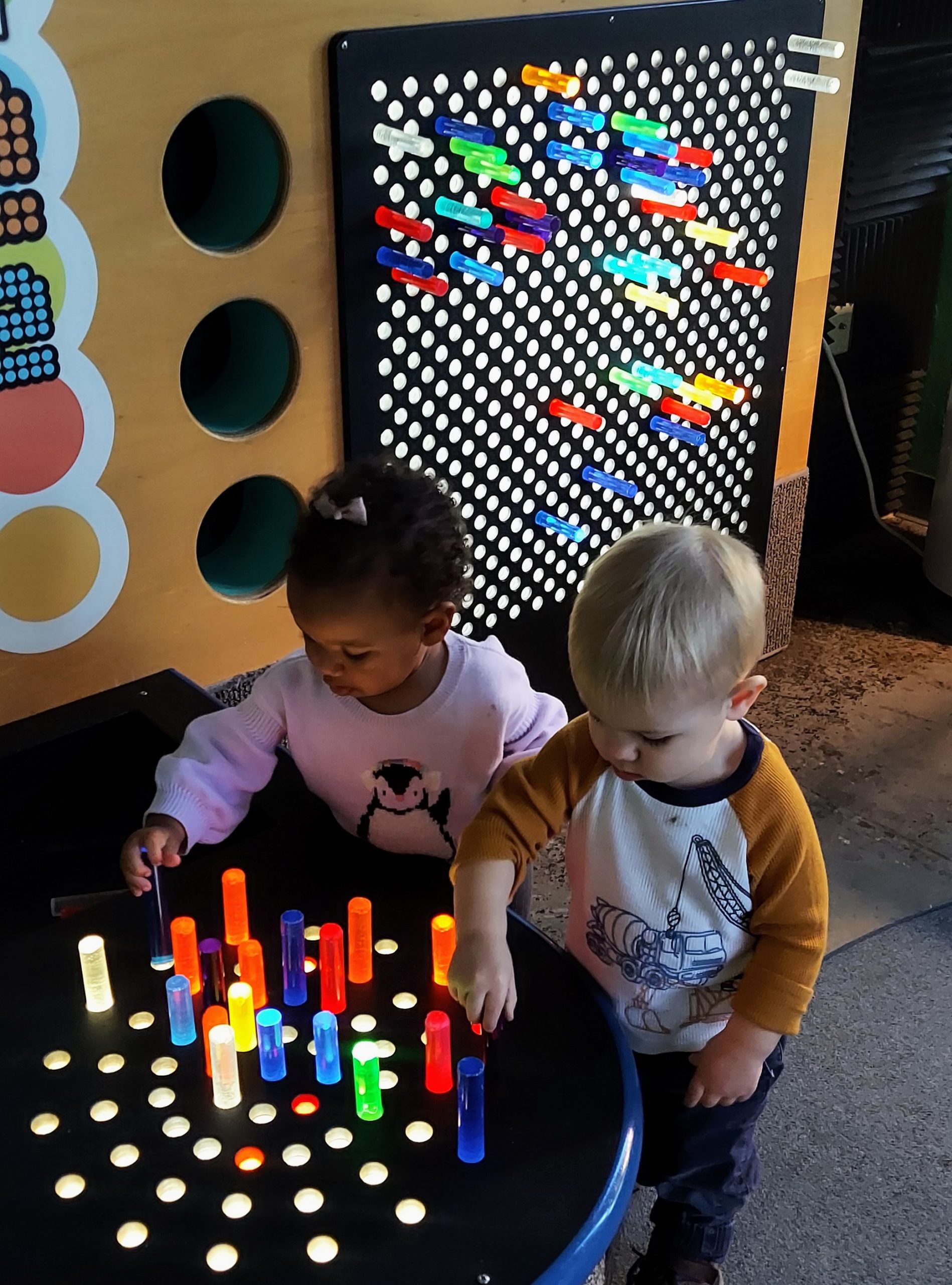Two toddlers reach for toy pieces of a giant Lite Brite shown in the background