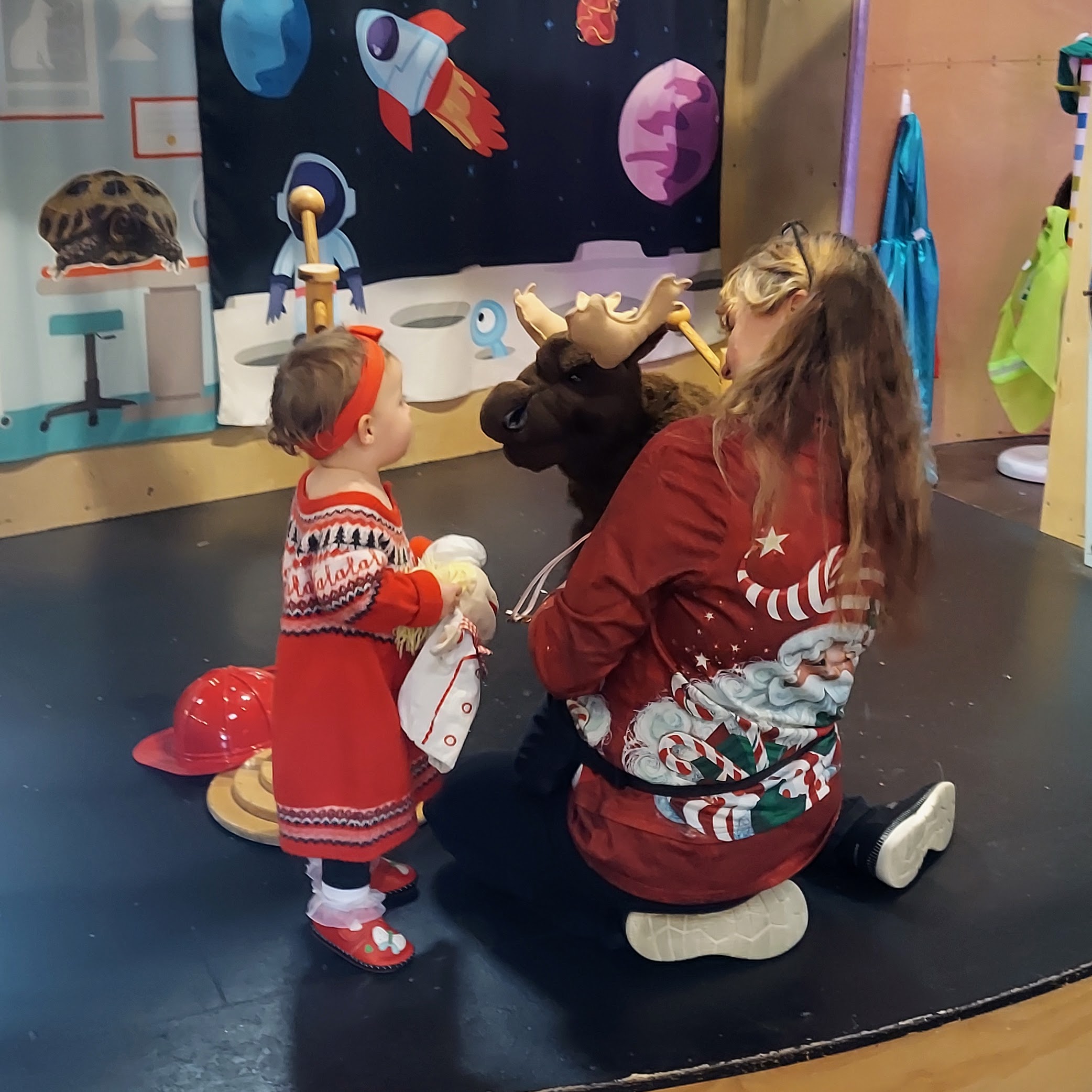 A woman manipulates a moose puppet, showing it to a young child in a red dress.