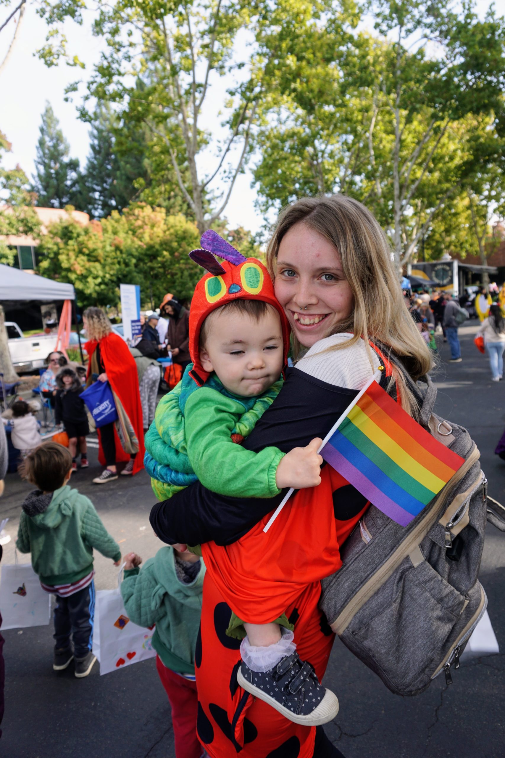 A mother smiles while holding her son, who is dressed as the Very Hungry Caterpillar and is holding a Pride flag