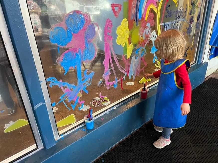A girl stands next to a clear wall covered in paintings, reaching for a paintbrush