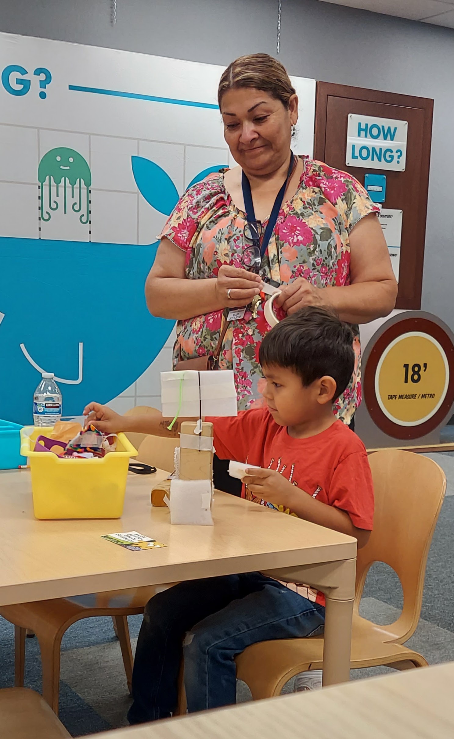 A boy builds a science experiment, reaching for another part from a yellow box, while a teacher watches over his shoulder, smiling