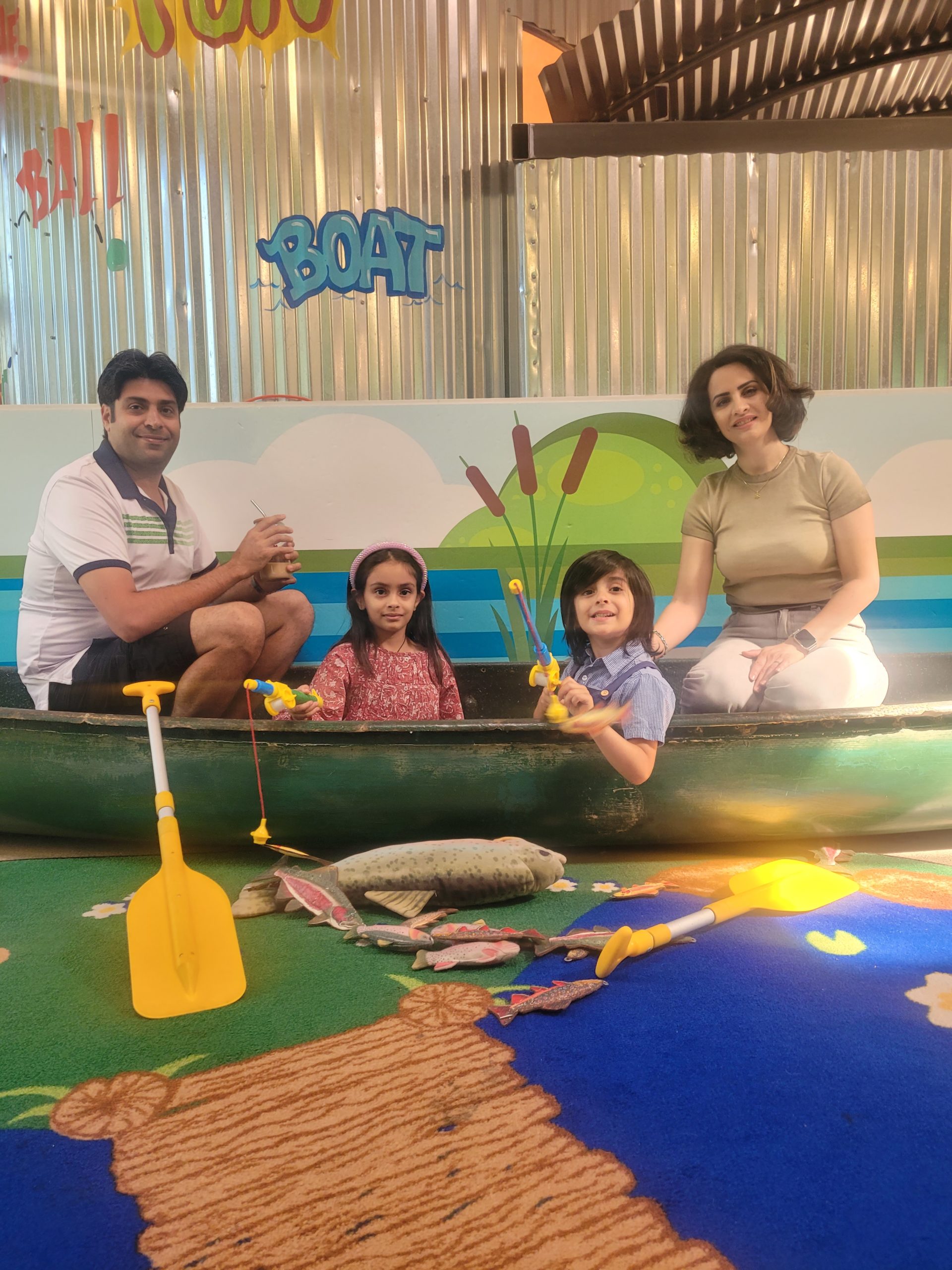 A family of 4 smiles in a play canoe with play fishing poles and a paddle